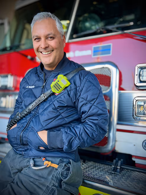 A firefighter in a quilted jacket sits on a fire truck, wearing a radio and with safety gear visible on his belt.