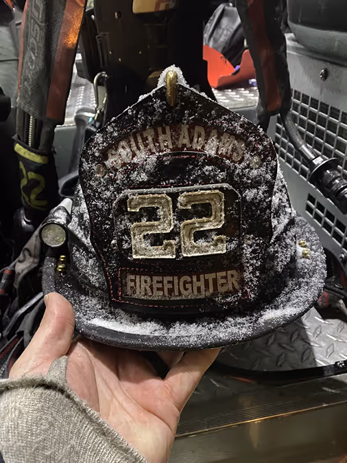 A firefighter's snowy helmet, labeled "South Adams 22," held in a hand against a dimly lit background.