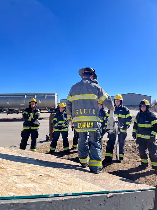A group of firefighters wearing uniforms and helmets gather outside for a training session under a clear blue sky.