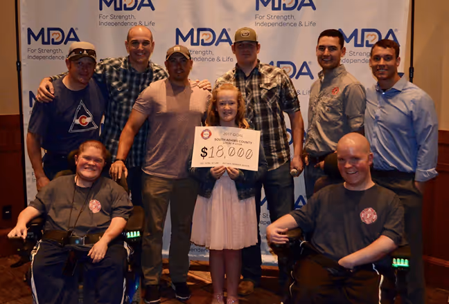 A group of ten individuals poses together, with one holding a sign that reads "$18,000," in front of a MDA backdrop.