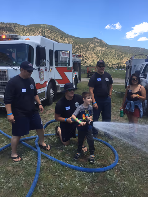 A child sprays water from a hose, assisted by firefighters near a fire truck in a scenic outdoor setting with mountains in the background.