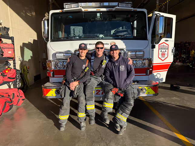 Three firefighters in gear sit on the front of a fire truck in a well-lit garage, showcasing a sense of camaraderie and teamwork.