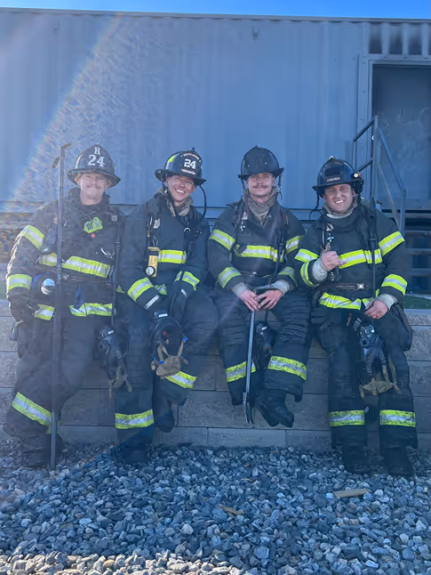 Four firefighters sit on a stone wall, wearing full gear with helmets, including yellow reflective stripes, against a gray container backdrop.