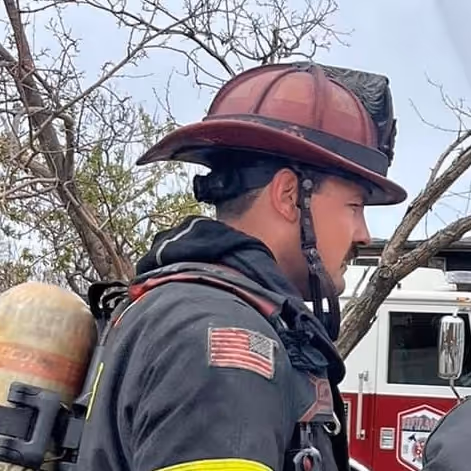 A firefighter in full gear stands with a helmet, oxygen tank, and an American flag patch, ready to respond to an emergency.
