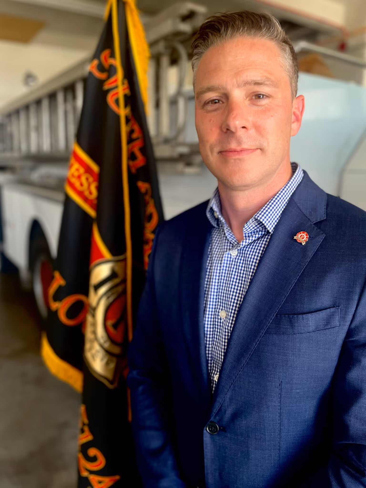A man in a dark suit stands beside a fire department flag, with a fire truck in the background, showcasing dedication and professionalism.
