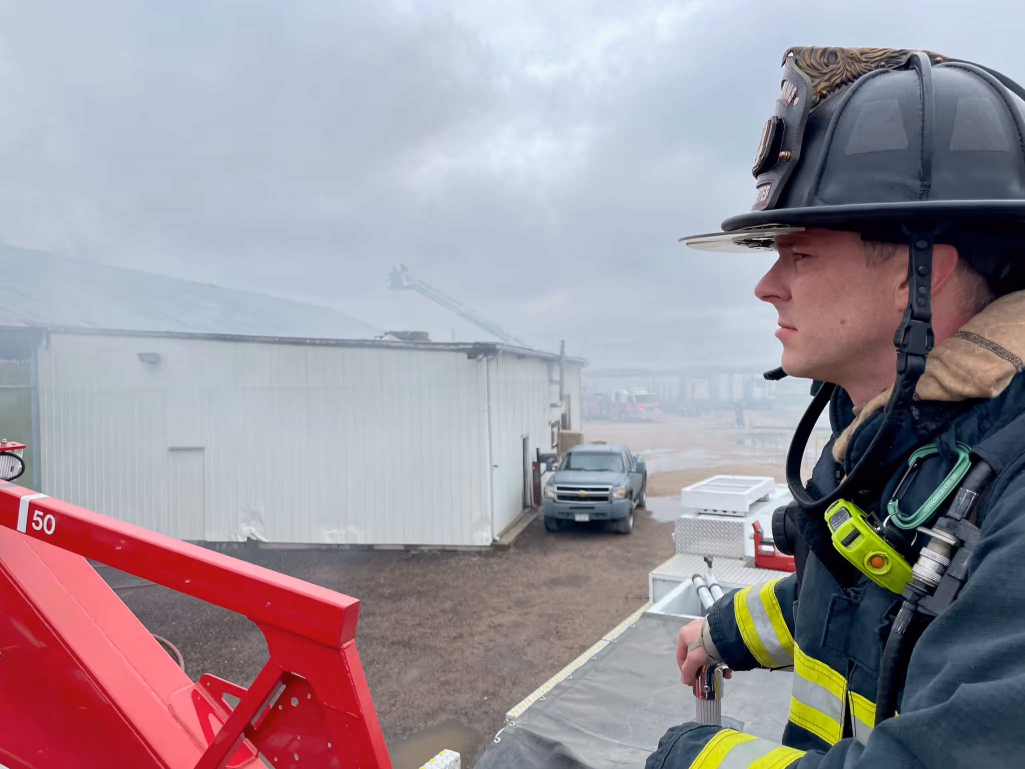 A firefighter in gear stands on a fire truck's ladder, observing a building shrouded in smoke on an overcast day.