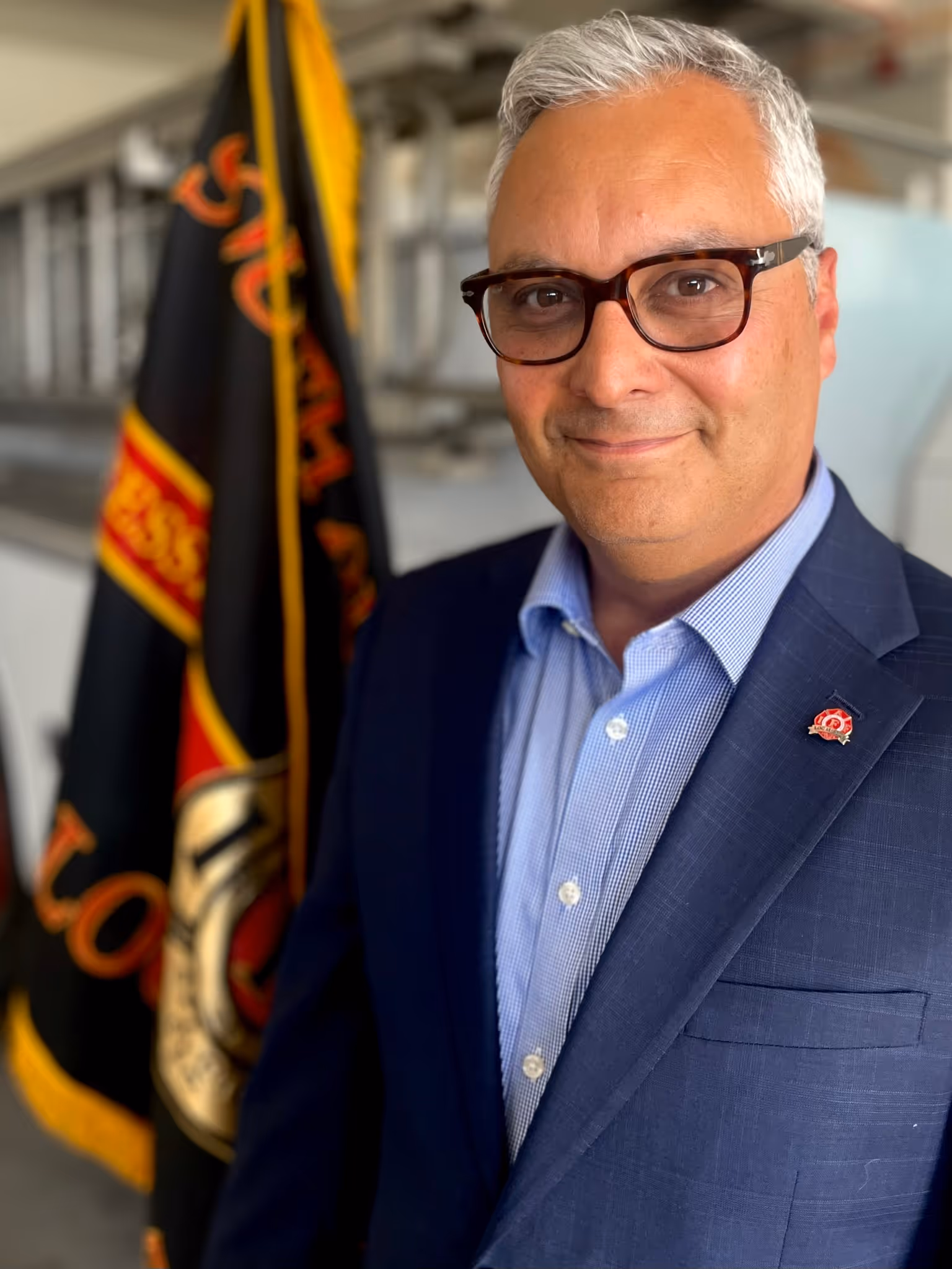 A man in a dark suit stands beside a fire department flag.