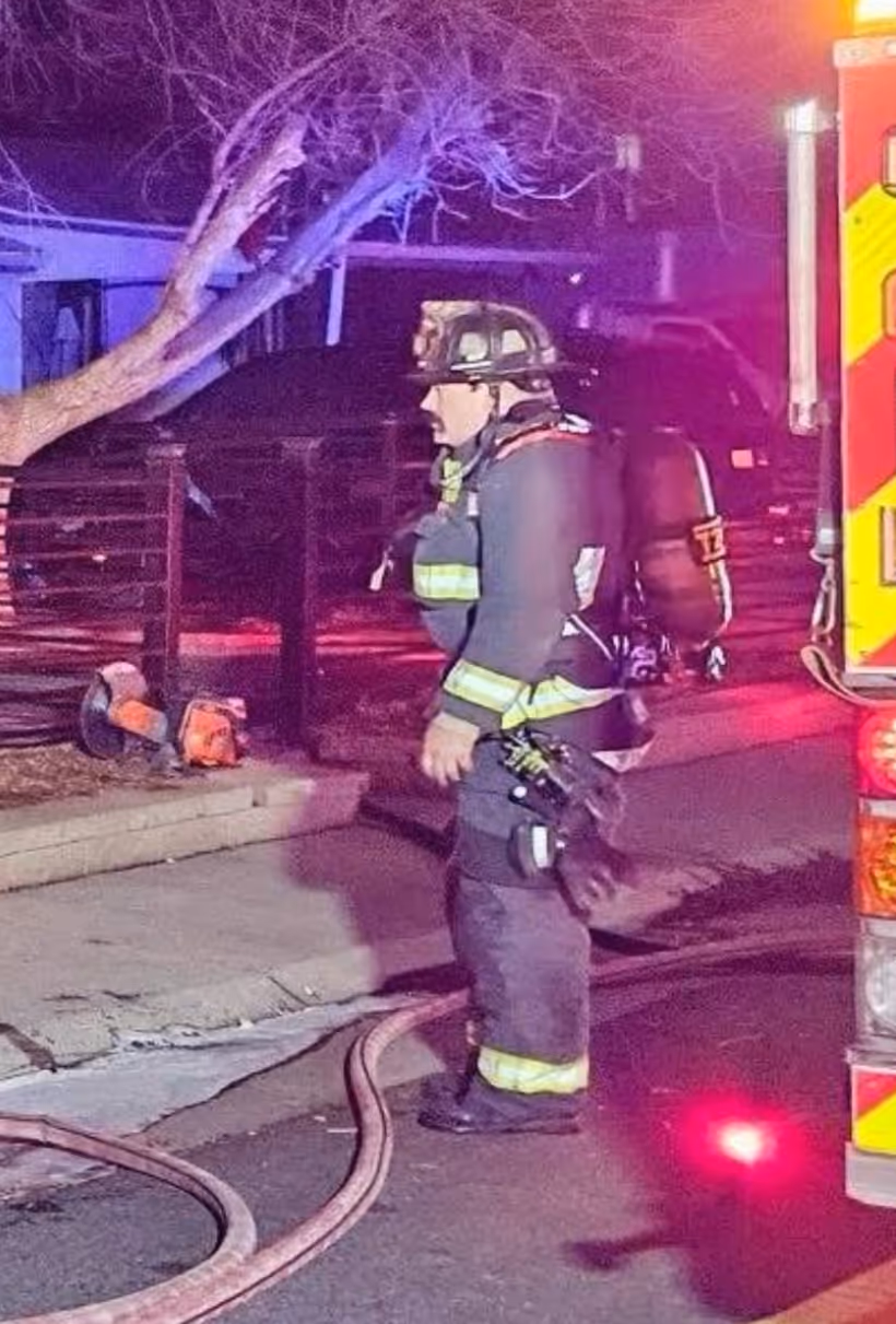 A firefighter in full gear stands near a fire truck, with hoses and emergency lights illuminating the scene amidst a nighttime setting.