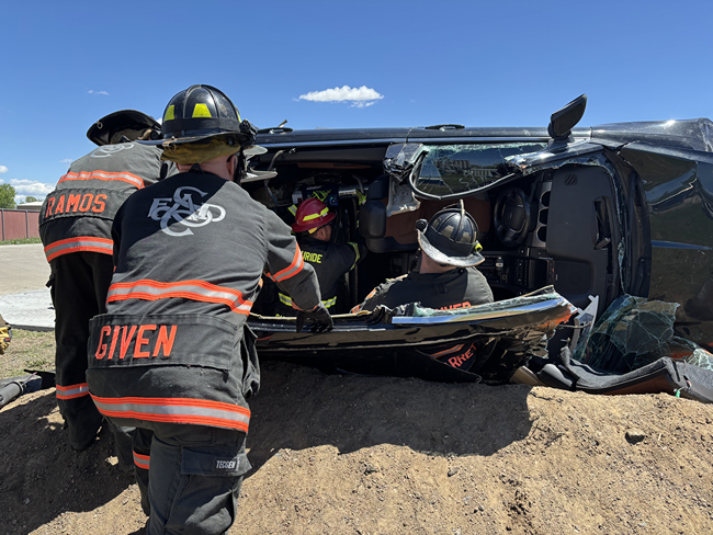 Firefighters in protective gear assist in rescuing victims from an overturned vehicle at an accident scene under a clear blue sky.