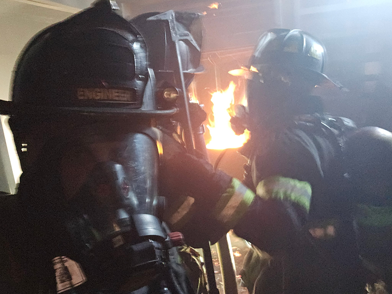 Two firefighters in protective gear and masks battle flames in a smoky environment, focused and determined in their efforts.