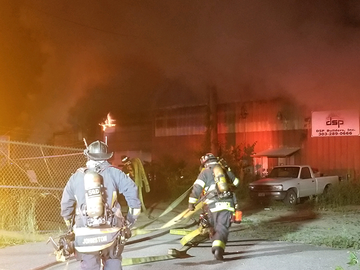 Two firefighters in gear maneuvering hoses toward a burning industrial building, engulfed in smoke and flames at night.