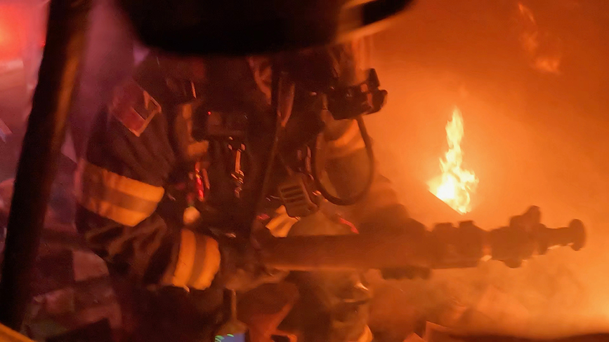 A firefighter in dark gear and helmet battles flames in a smoky environment, aiming a hose towards a bright fire.
