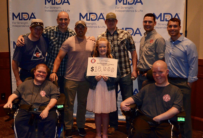A group of individuals poses together, holding a large check for $18,000, against an MDA banner for a fundraising event.