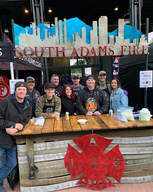 A group of firefighters stands behind a wooden booth with a "South Adams Fire" sign