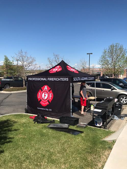 A firefighter-themed tent set up in a parking lot, with a person preparing food at a grill under clear blue skies.