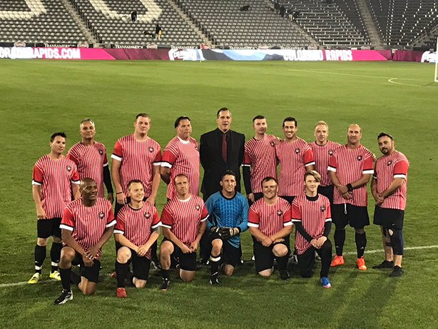 A soccer team poses for a group photo on a field, wearing red striped jerseys, with a goalpost and empty stadium seats in the background.