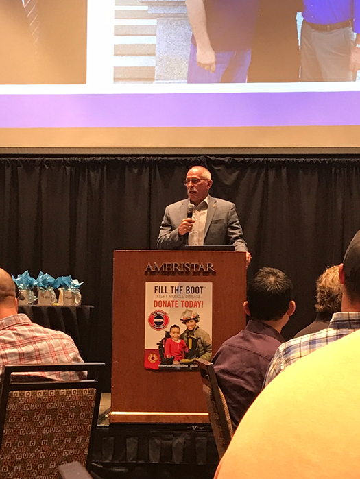 A speaker stands at a podium in front of an audience, promoting a charity event with a "Fill the Boot" sign.