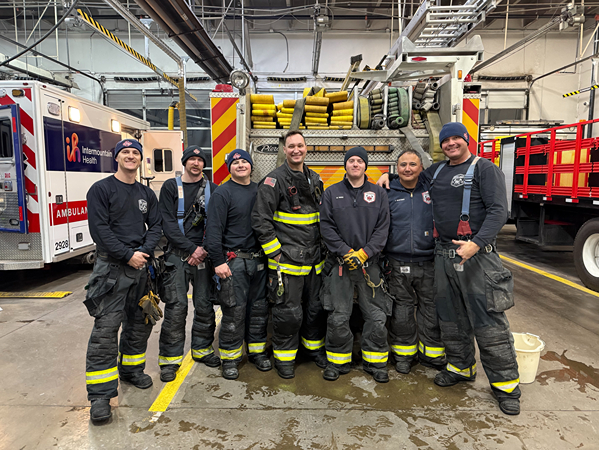 A group of firefighters in protective gear stands together in a fire station, with a fire truck and ambulance in the background.