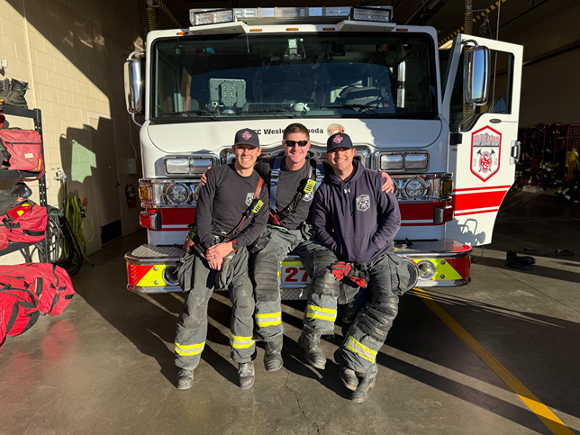 Three firefighters seated on the front of a fire truck, dressed in turnout gear, with equipment and gear bags in the background.