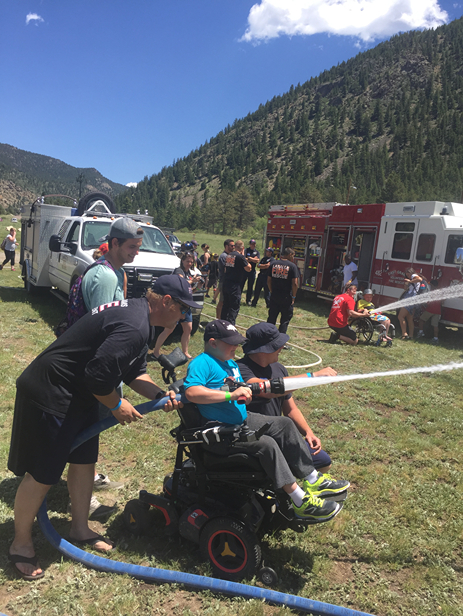 A group of people participate in a water spray activity, with emergency vehicles and mountains in the background, under a clear blue sky.