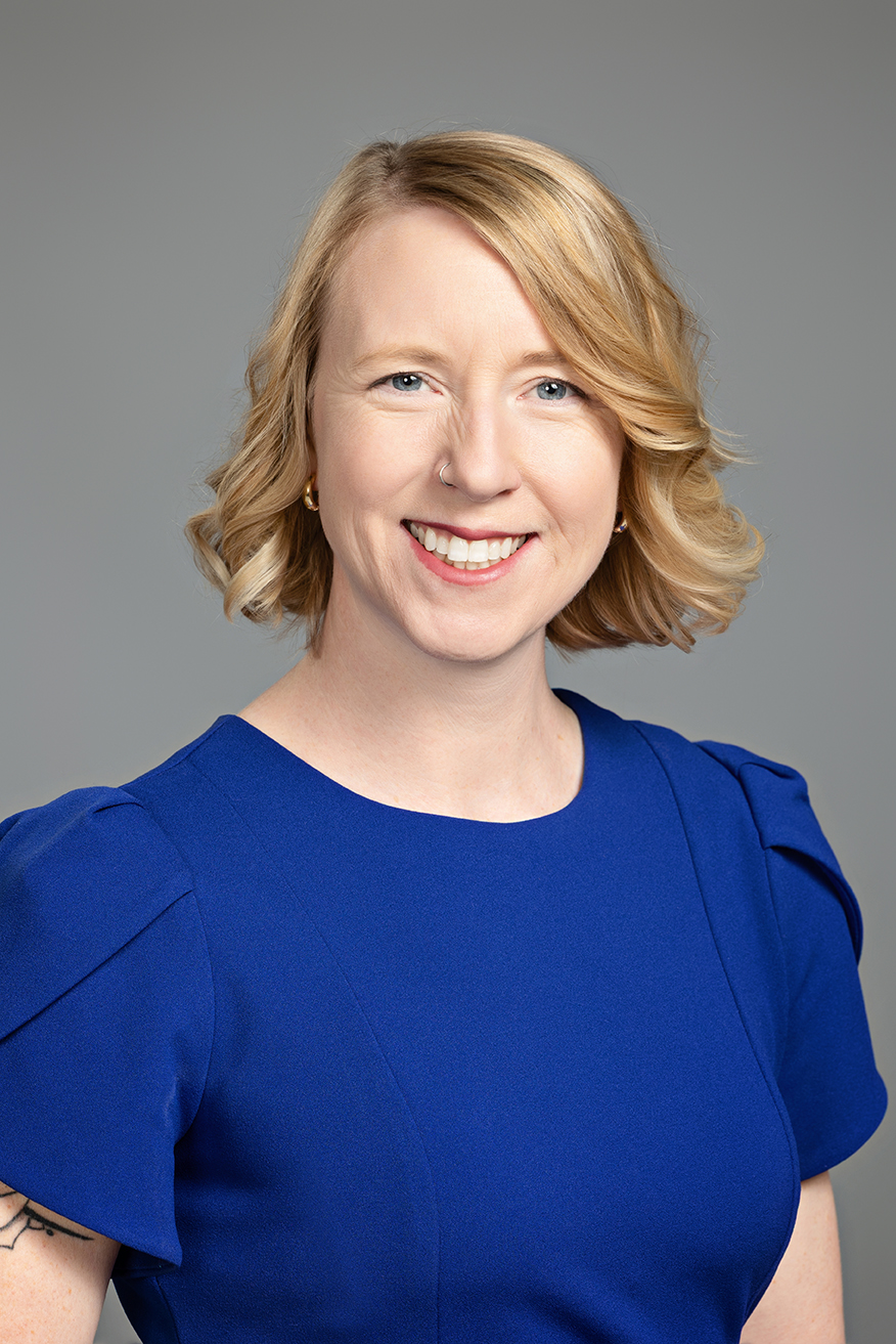 Smiling woman with short curly blonde hair wearing a blue dress, posed for a professional headshot against a grey background