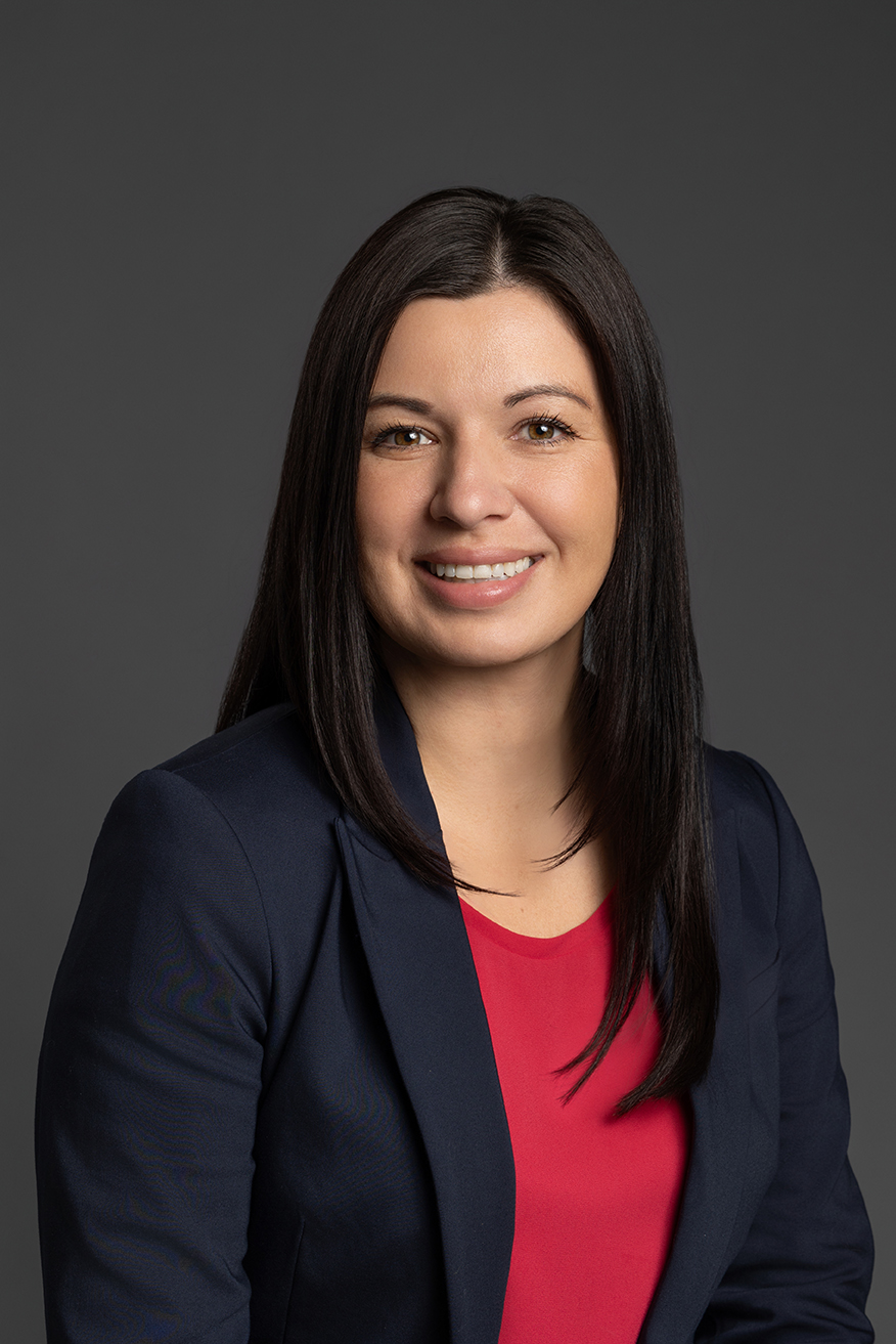 Smiling professional woman with straight dark hair wearing a navy blazer and red top, posed against a grey background