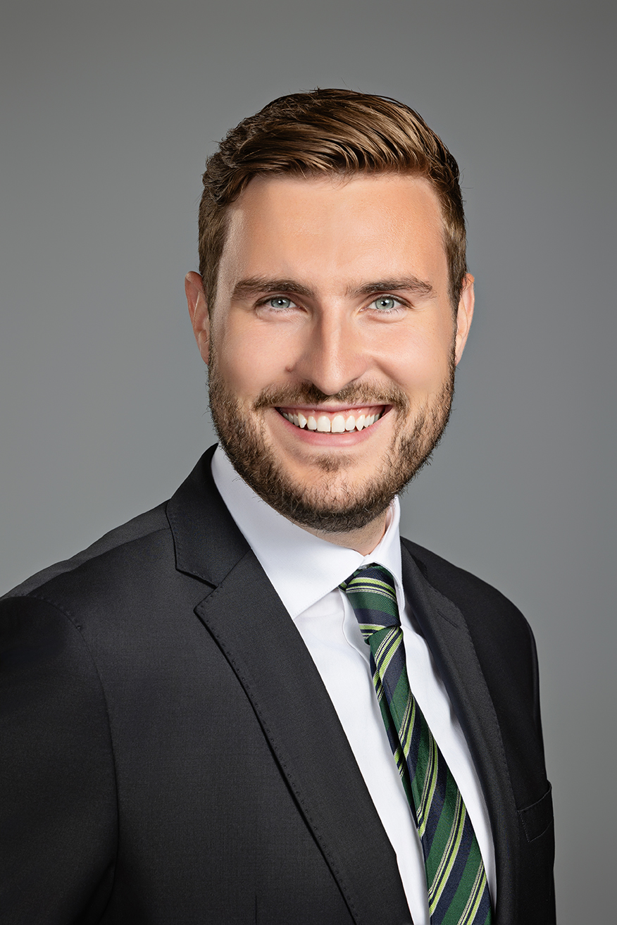 Professional headshot of a smiling man in a dark suit with a green striped tie and white shirt, posed against a grey background