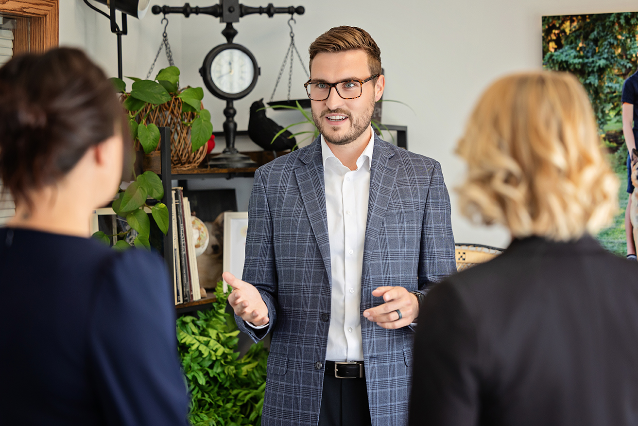 Male business consultant in a checkered blazer speaking to two team members in a decorated office setting