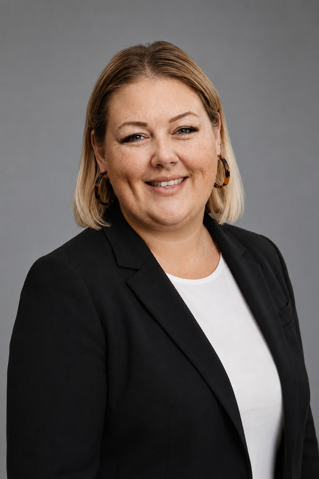 Portrait of Michele smiling, wearing a black blazer over a white top against a grey background.