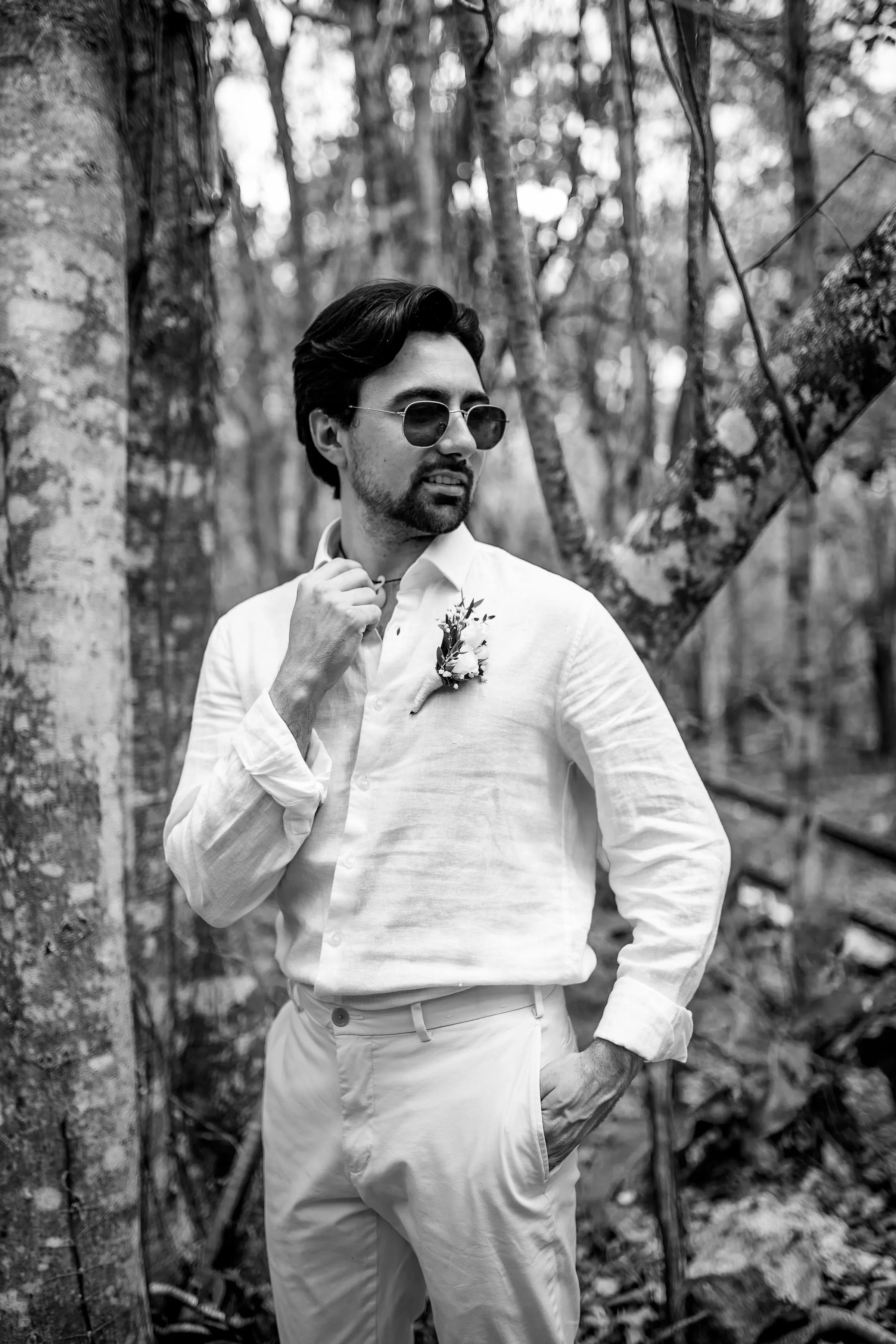 Black and white portrait of a man wearing sunglasses and a light-colored shirt with a boutonnière, standing confidently in a forest with one hand in his pocket and the other adjusting his collar.