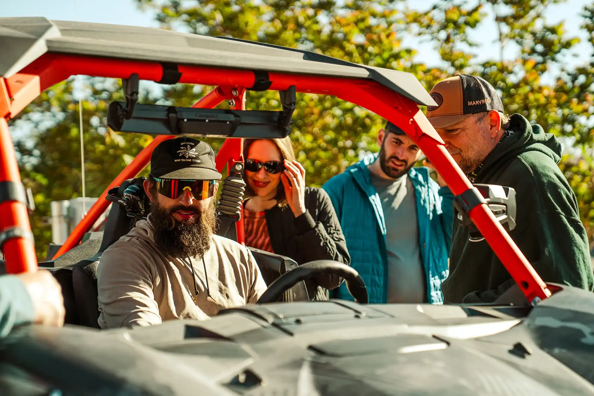 Four adults, one seated in an off-road vehicle wearing a black cap and sunglasses, with three others standing outside nearby on a sunny day.