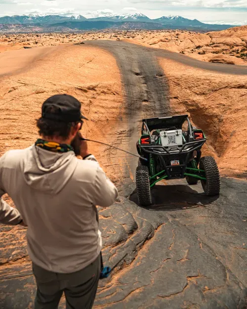 Man guiding an off-road vehicle climbing a steep rocky hill with mountains in the background.
