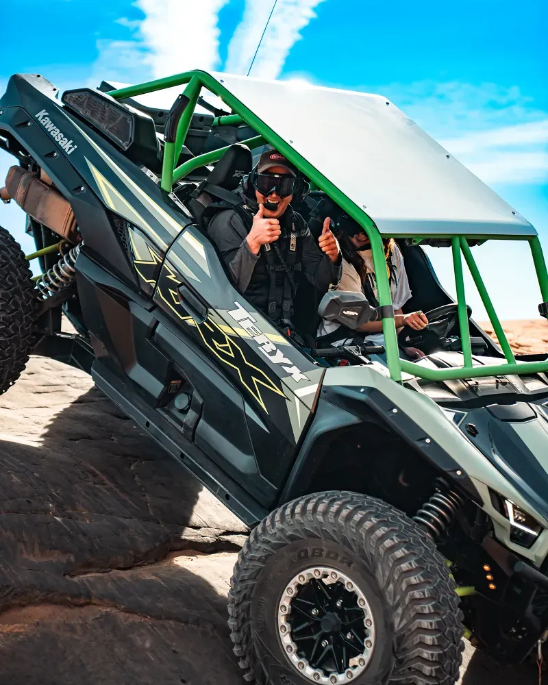 Two people riding a green and black Kawasaki Teryx off-road vehicle on rocky terrain under a blue sky.