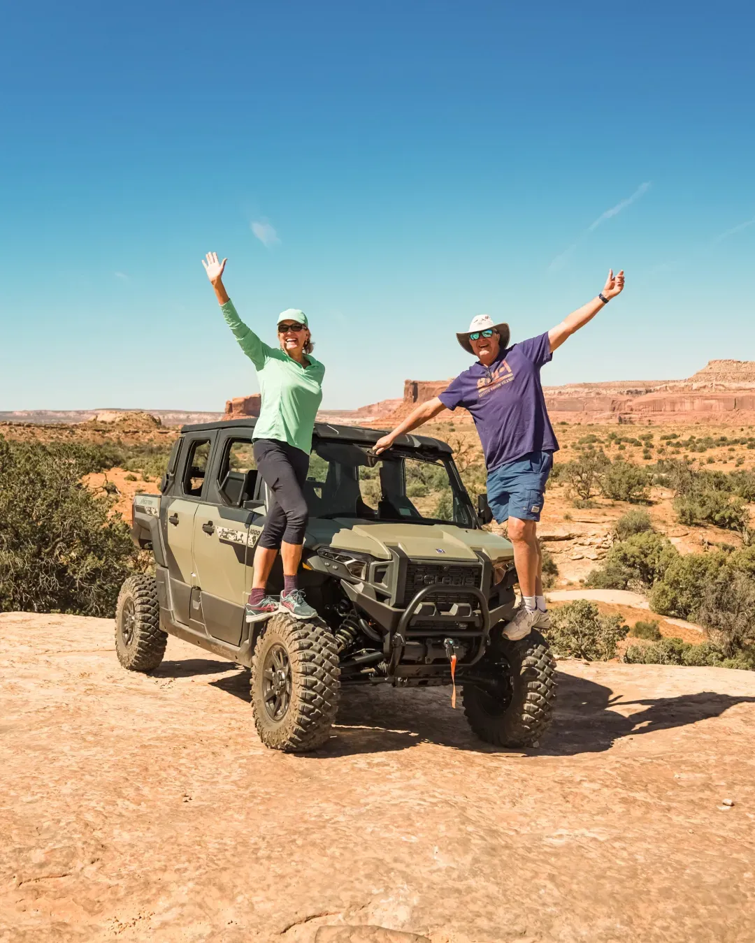 Two people standing on the front of a Polaris off-road vehicle, smiling and waving in a desert landscape with clear blue sky.