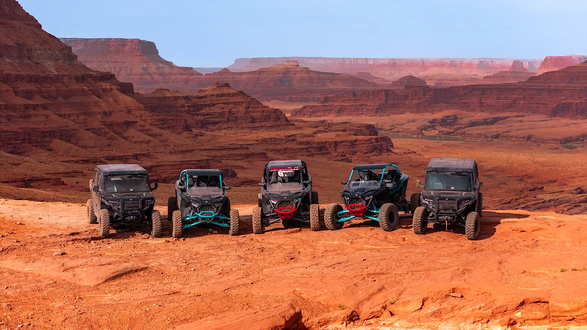 Five off-road UTVs lined up on red dirt plateau with layered red rock canyon landscape in the background.