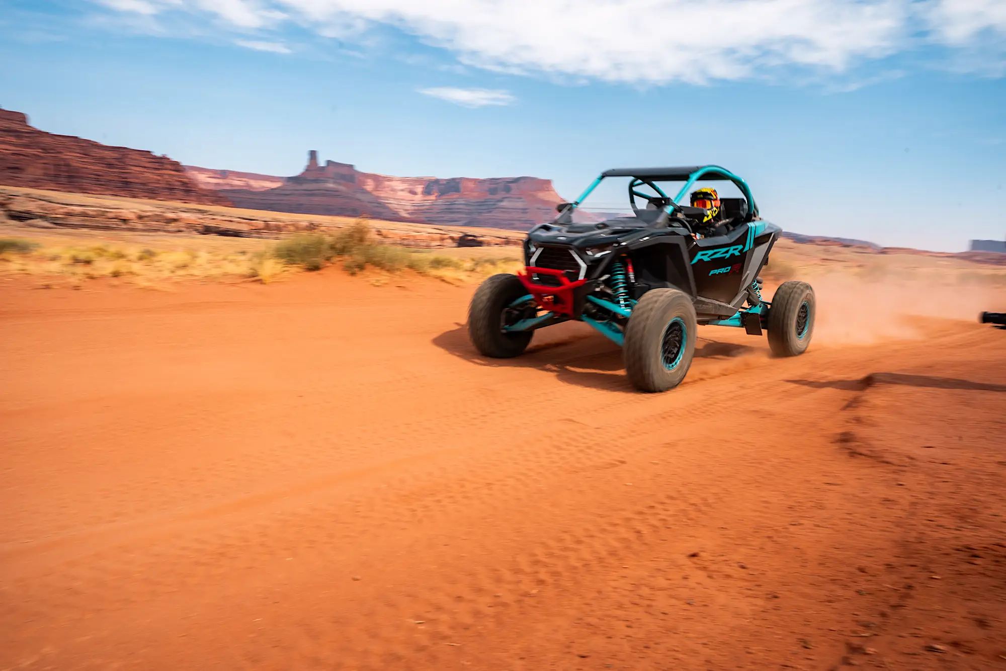 Black and turquoise off-road dune buggy speeding across red desert sand with rocky mesas in the background.