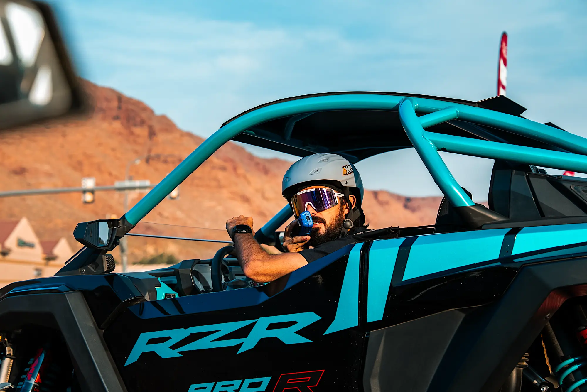 Man wearing helmet and goggles driving a blue and black RZR off-road vehicle in a desert landscape.