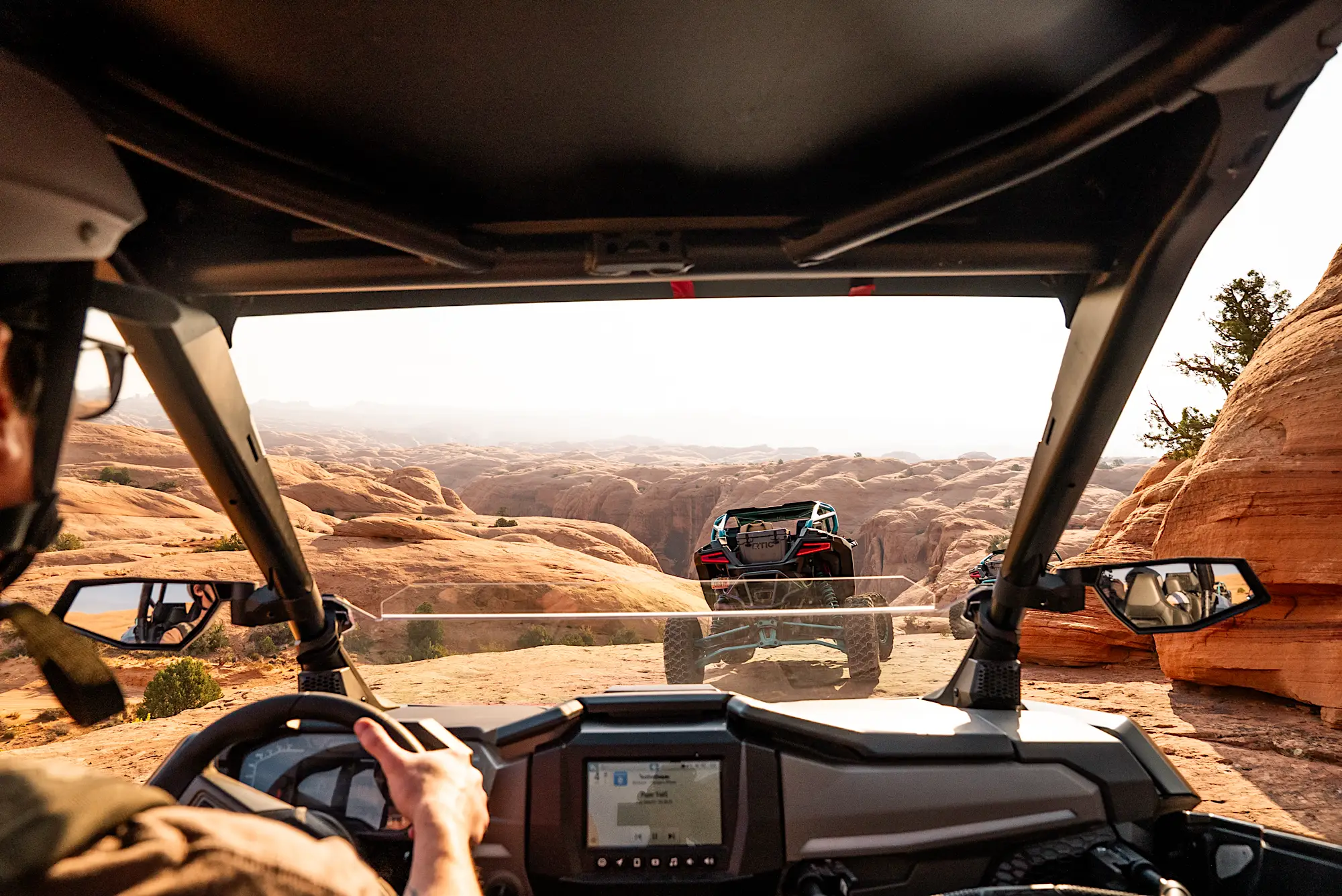View from inside an off-road vehicle driving on a rocky desert trail with another off-road vehicle ahead among red rock formations.