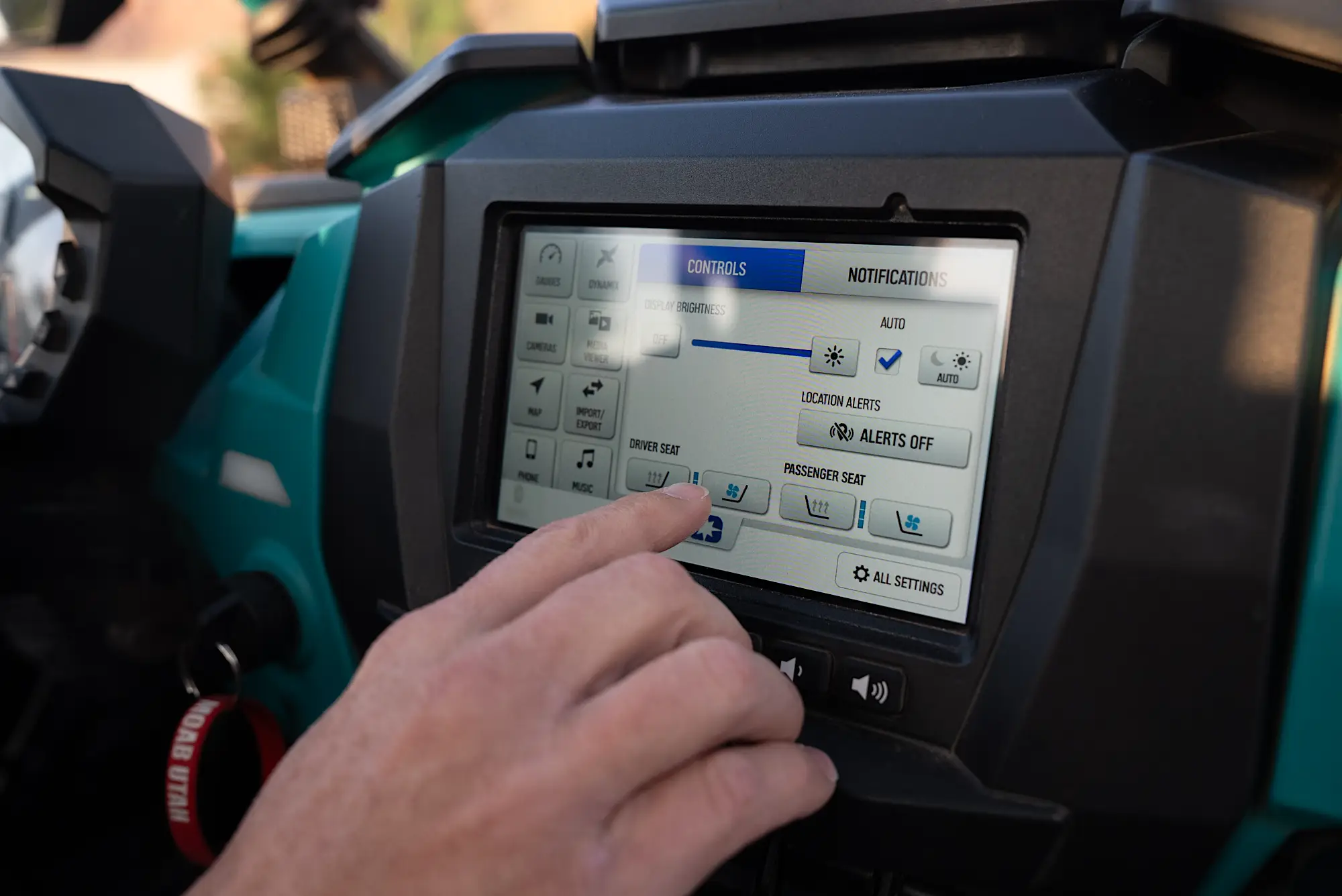 Hand adjusting the touchscreen controls of a vehicle, showing brightness, location alerts, and seat heating and cooling options.