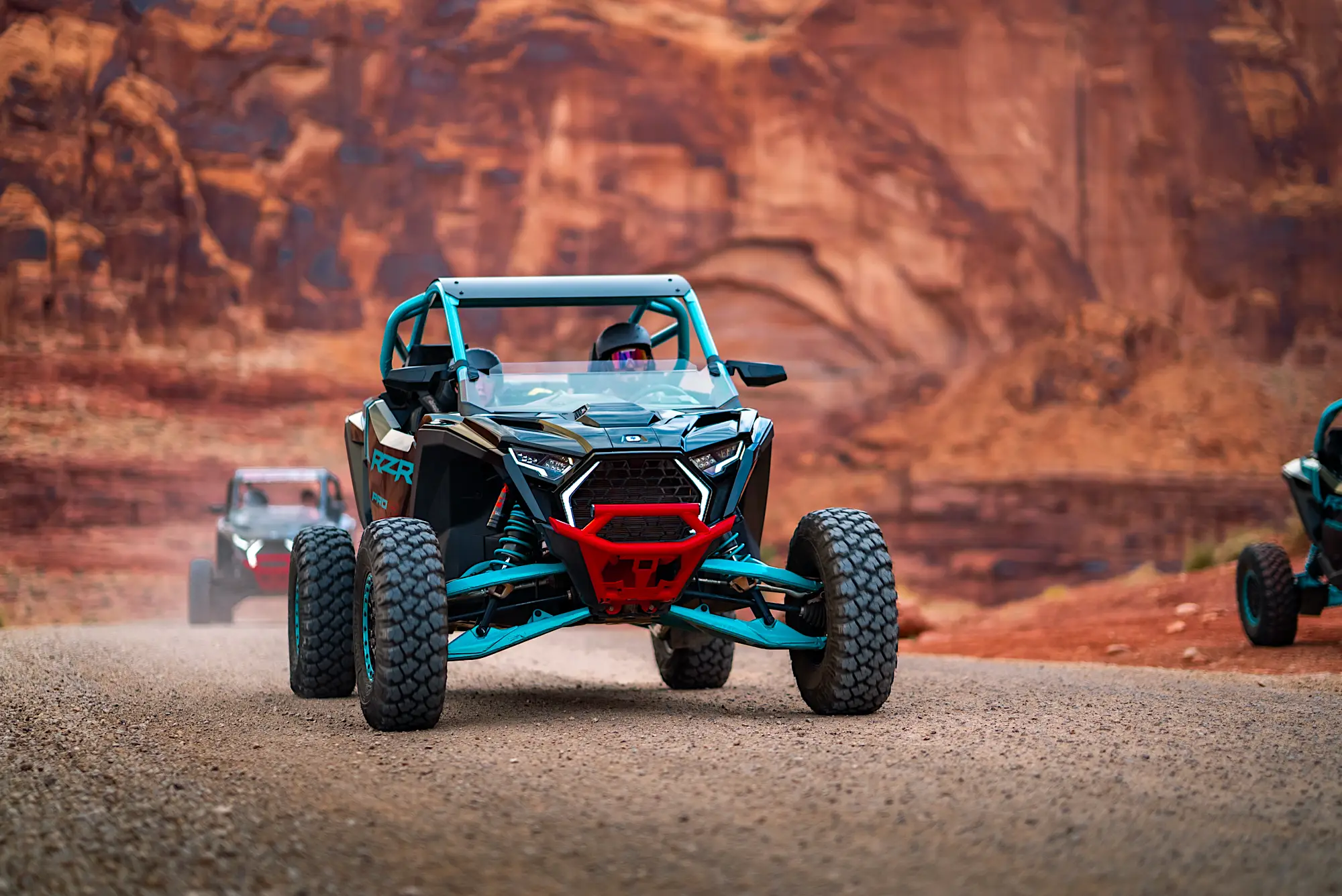 Two off-road dune buggies driving on a dirt road with red rock formations in the background.