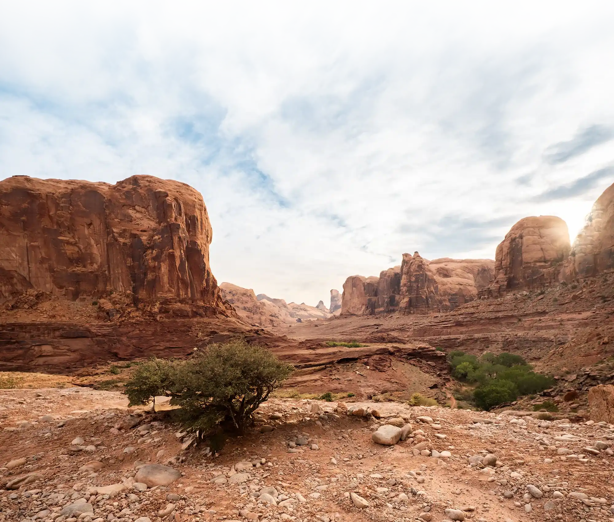 Rocky desert landscape with large red sandstone cliffs under a partly cloudy sky and sparse vegetation.