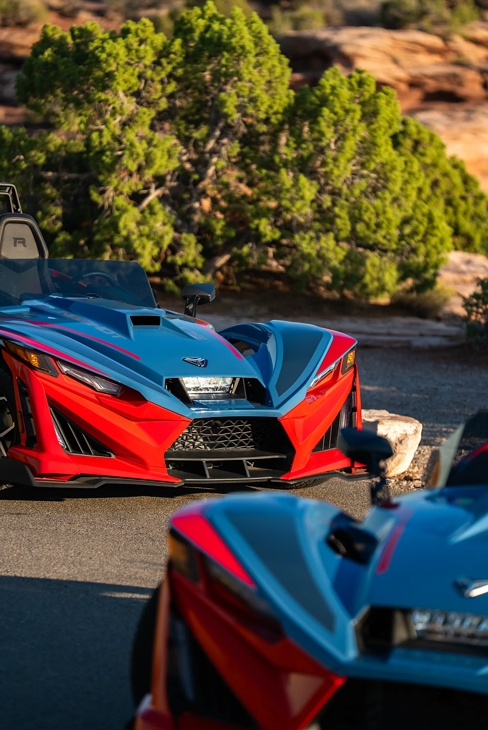 Two sporty red and blue three-wheeled vehicles parked on a road with green shrubs and rocks in the background.