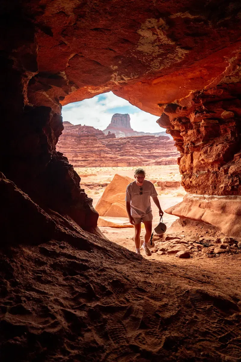 Person walking out of a red rock cave with desert landscape and rock formations in the background.