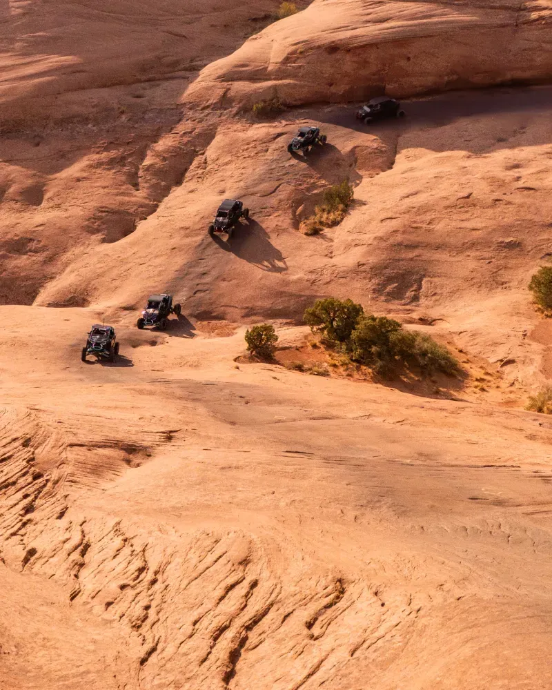 Aerial view of five off-road vehicles driving on a winding rocky terrain with sparse shrubs.