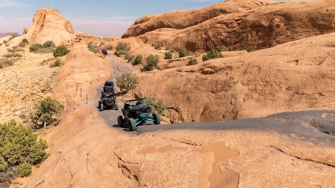 A group of off-road vehicles driving up a rocky, desert hill surrounded by red rock formations and sparse greenery.
