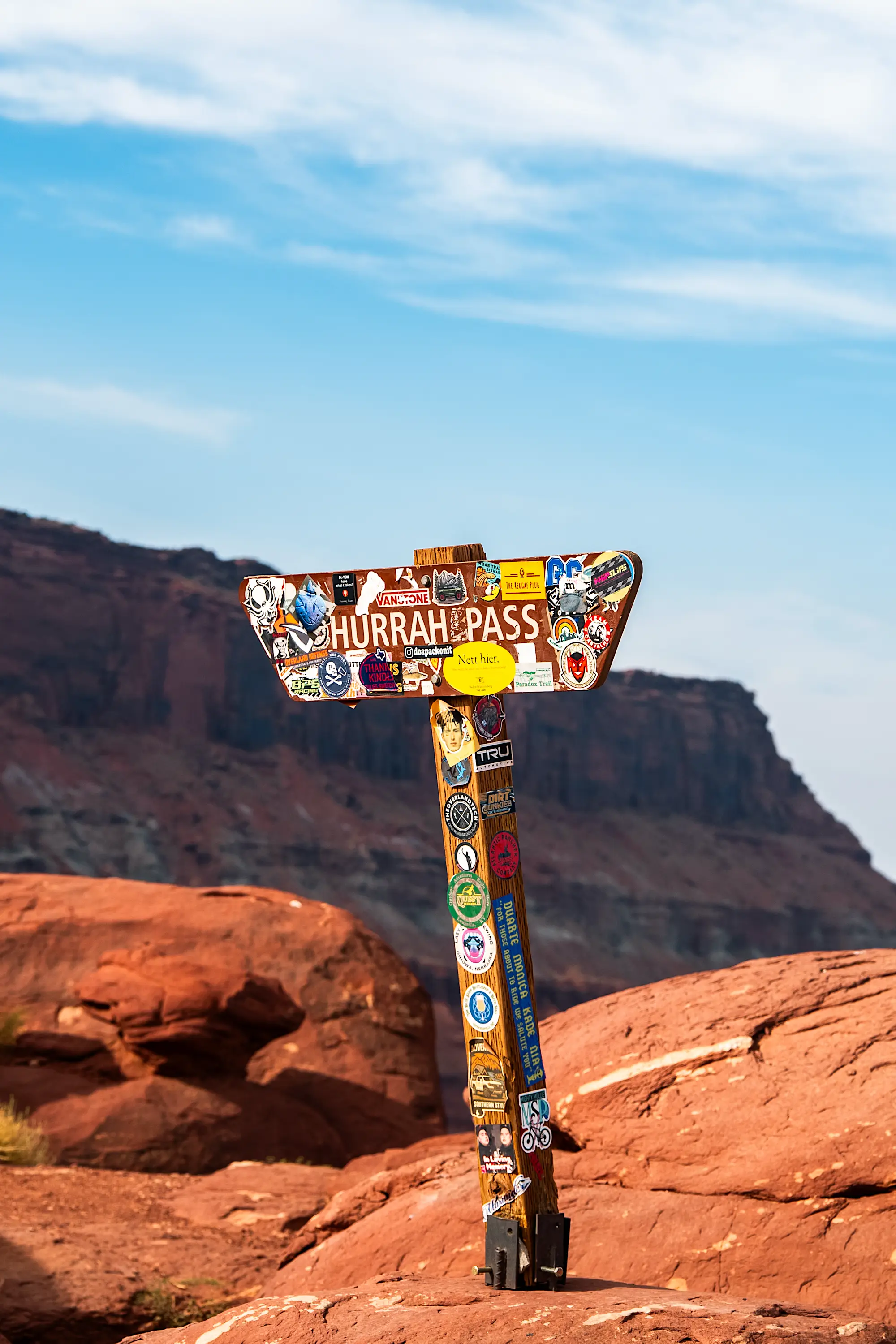 Wooden Hurrah Pass sign covered with colorful stickers against a rocky desert landscape under a partly cloudy blue sky.