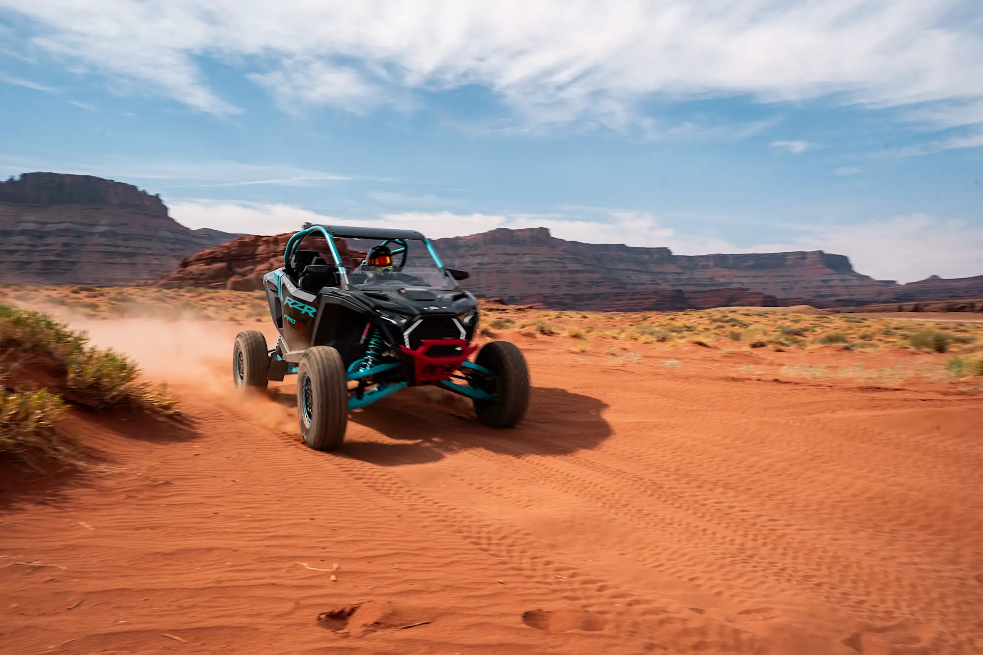 Black and blue off-road RZR vehicle kicking up dust on a red dirt trail in a desert landscape with rocky cliffs in the background.