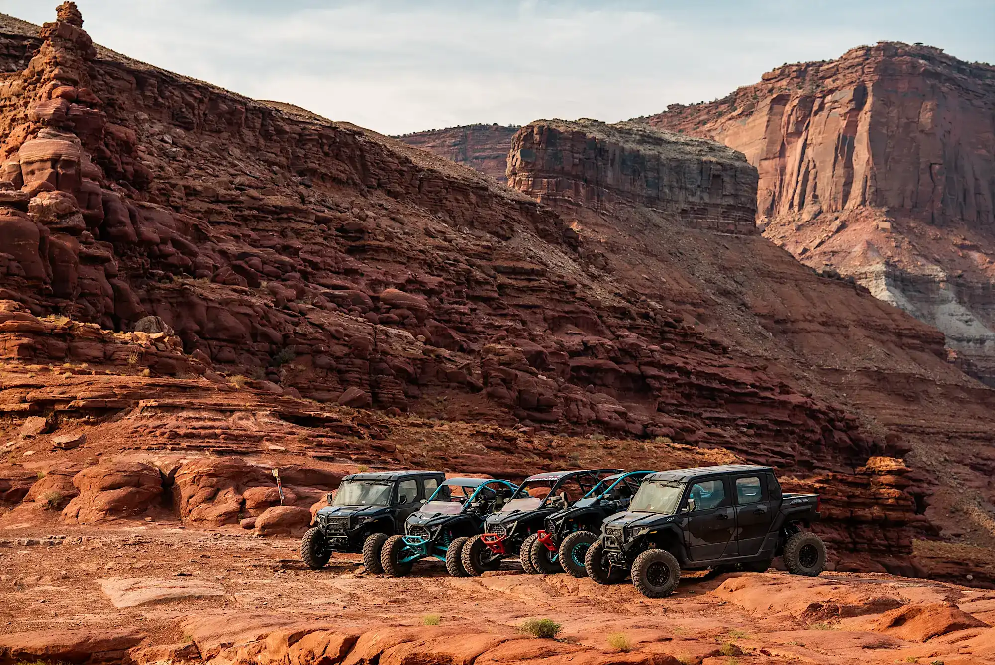Five off-road vehicles parked on red rocky terrain with large canyon cliffs in the background.