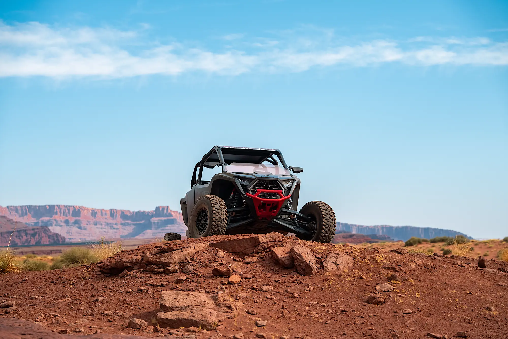 All-terrain vehicle parked on a rocky red dirt hill with desert cliffs in the background under a blue sky.