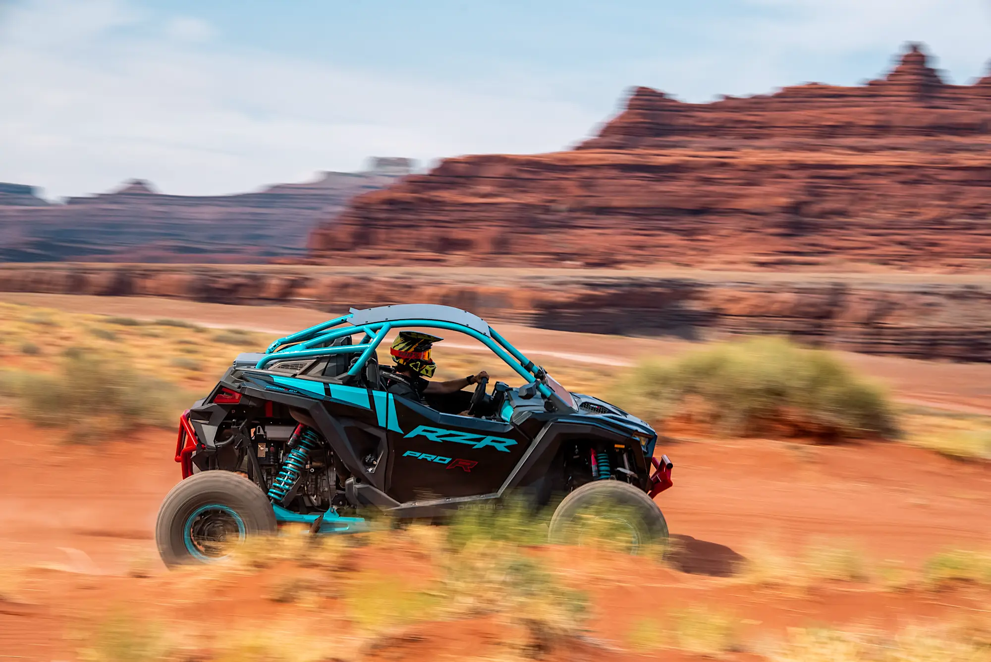 Person wearing helmet driving a blue Polaris RZR off-road vehicle on red dirt with rocky desert landscape in the background.